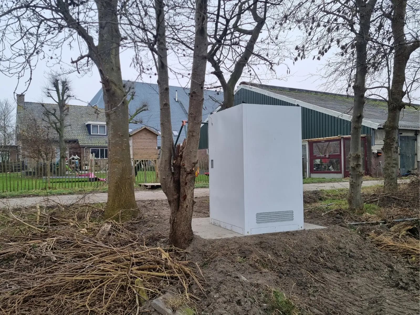 Witte batterijkast in buitenopstelling tussen de bomen op het erf van een melkveehouderij, met de stal en het woonhuis op de achtergrond