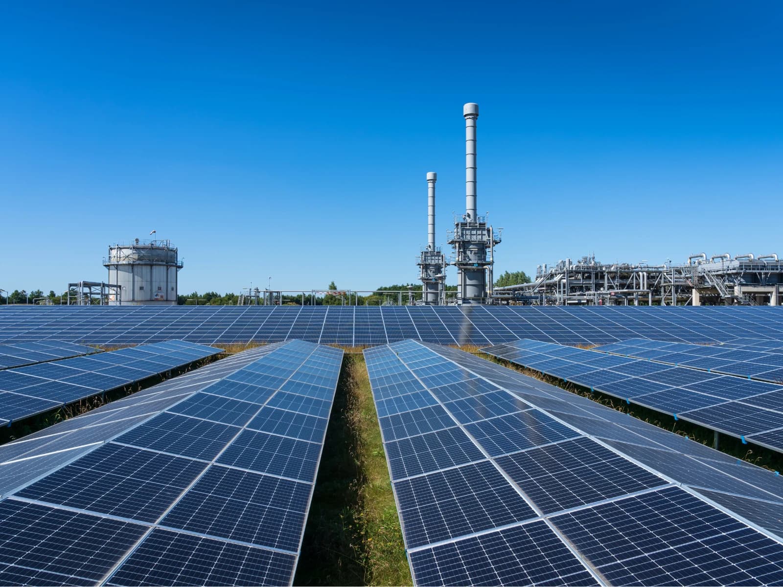 Zonnepark met rijen zonnepanelen voor een industrieel complex onder een strakblauwe lucht